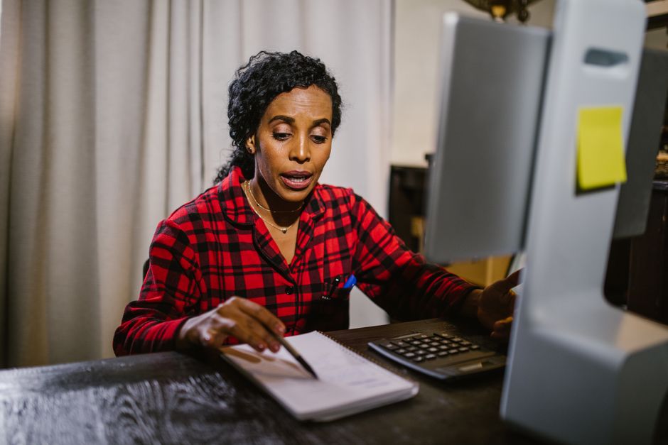 Mujer afroamericana con camisa a cuadros trabajando en el ordenador en casa con un cuaderno y una calculadora.