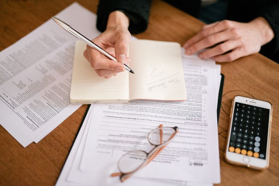 Primer plano de una persona escribiendo en un cuaderno con documentos y una calculadora, gestionando finanzas.
